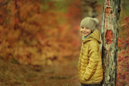 little boy in a yellow jacket and knit cap stands near a tree in autumn forest