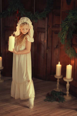 against the backdrop of pine needles Pretty blond girl in a pink dress and a cap with a candleの写真素材