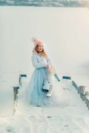 snowy winter wooden bridge near the frozen river is a cute girl in a magnificent dress with skatesの写真素材