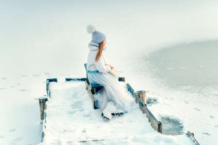 snowy winter wooden bridge near the frozen river is a cute girl in a magnificent dress with skatesの写真素材