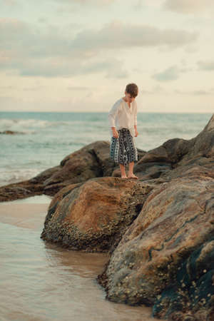 A hot day in the ocean, a cute, barefooted boy stands on large stonesの写真素材