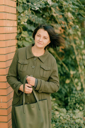 girl in green coat with green bag stands near a brick wall
