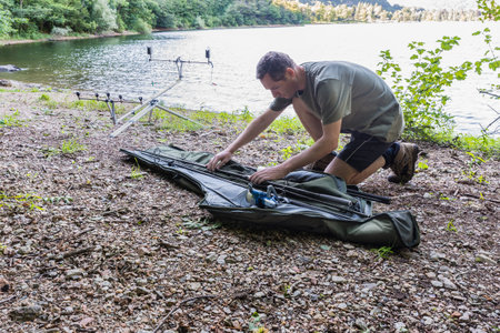Fishing adventures, carp fishing. Fisherman on the shore of a lake is taking fishing rods from a rod holdallの写真素材