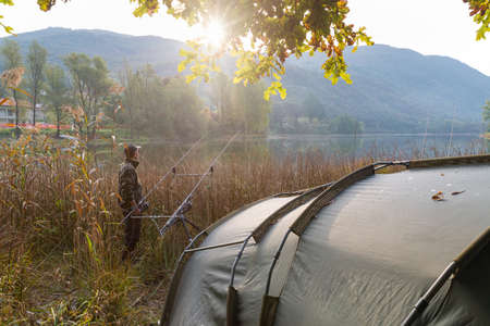 Fishing adventures, carp fishing in autumn. Camping on the lake at sunrise; sun rising from the mountain and fisherman with camouflage fleece on the shore, hidden behind the reedsの写真素材