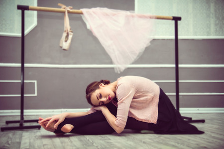 young beautiful graceful ballerina resting in ballet class sitting on the floor, ballet shoes, a pink skirt and ballet barre in the backgroundの写真素材