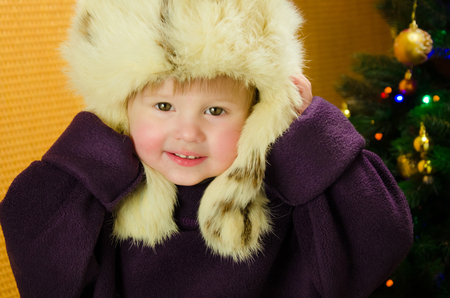 Portrait of two years old pretty little girl in white, russian, fur hat near a Christmas treeの写真素材