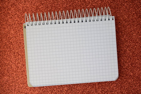 A notebook with an open blank sheet for writing lies on a red shiny surface.