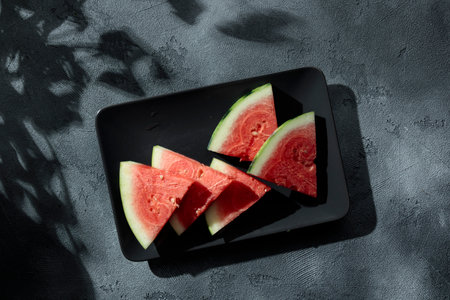 Slices of juicy watermelon on the black plate on the concrete background with summer sun shadows. Copy space. Top view.の写真素材