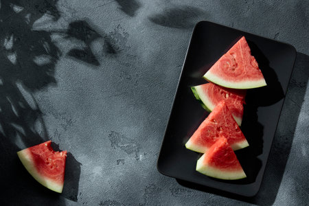 Slices of juicy watermelon on the black plate on the concrete background with summer sun shadows. Copy space. Top view.の写真素材