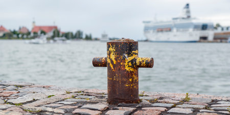 Old rusty steel mooring bollard pole on a pier and a sea and ferry on the background, on the city,...