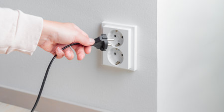 A woman's hand unplugging a power cord from an electrical outlet on a white wall. Careful resource use, the rising cost of electricity and the need for mindful consumption, safety and responsible energy management in everyday lifeの写真素材