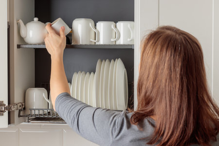 A young woman puts a mug in a dish-drying rack neatly integrated into a wall cabinet in a modern kitchen. The rack holds white ceramic kitchenware, plates and mugs, all arranged orderly. Well-organized aesthetic of simplicity and efficiencyの写真素材
