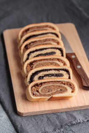 Bejgli, home-baked roll, traditional Hungarian, Eastern European Christmas and Easter sweet bread in slices stuffed with ground walnuts and poppy seed filling on wooden cut board on a gray background. Vertical top view on a gray backgroundの写真素材