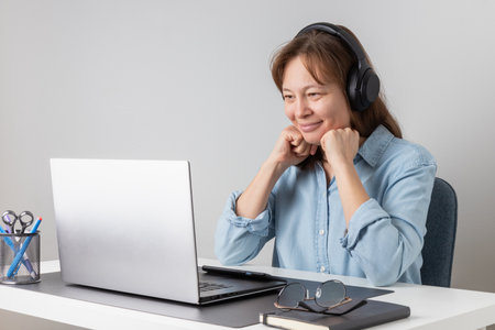 Smiling freelance professional woman wearing big headphones listening and watching video, online conference, video meeting, webinar or studying at the web course on laptopの写真素材