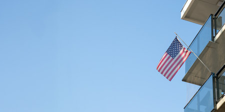 A balcony of a residential building with an American flag waving gently against a clear blue sky. A...