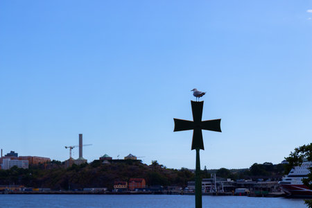 A seagull perched on a metal cross against the backdrop of the sea and sky, captured on a calm summer day in Stockholn, the Swedish capitalの写真素材