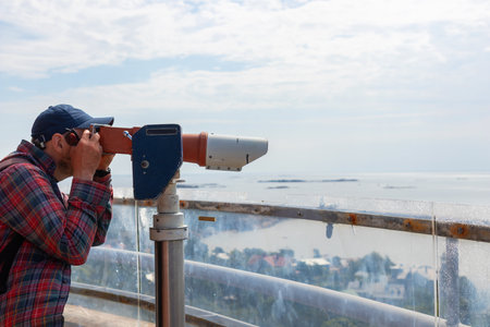 A young male tourist looks through a viewing binocular on an observation deck of Hanko Water Tower,...
