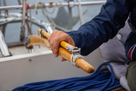 Skilled young man steering a sailboat with wooden tiller on the open waters of the Gulf of Finland. Good sailing experience, maritime adventure, summer leisure, connection to the seaの写真素材