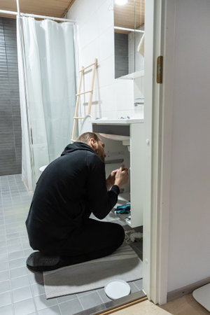 Handyman installing a new faucet and plumbing lines under a bathroom sink in a modern bathroom with tiled floor. Home renovation scene showing repair, maintenance, and proper installation workの写真素材