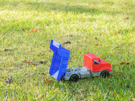 Red and gray dump truck toy with blue tipper bed on a sunlit green lawn. Durable plastic, safe for indoor and outdoor play, surrounded by scattered autumn leavesの写真素材