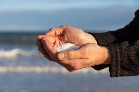 Cupped hands holding natural white sea salt crystals with blue ocean waves and clear sky in the background, symbolizing purity, nature, and coastal livingの写真素材