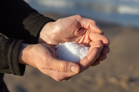 Cupped hands holding natural white sea salt crystals with blue ocean waves and clear sky in the background, symbolizing purity, nature, and coastal livingの写真素材