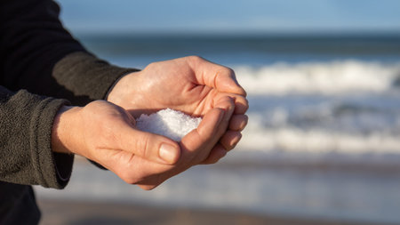 Cupped hands holding natural white sea salt crystals with blue ocean waves and clear sky in the background, symbolizing purity, nature, and coastal livingの写真素材
