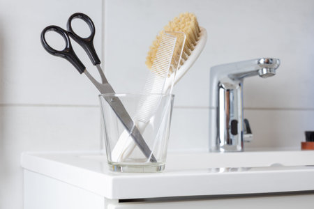 Hair scissors and comb in a glass next to a bathroom faucet on the sink in the modern minimalistic...