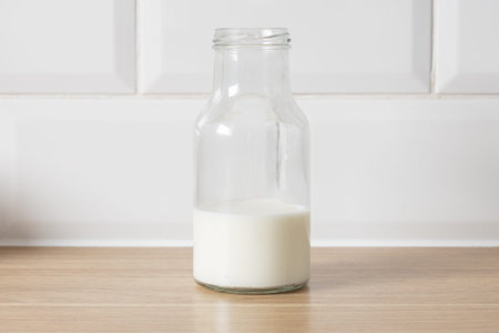A simple glass bottle of milk sits on a wooden countertop, with a clean white tiled background.