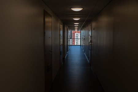The long, dimly lit corridor of a modern residential building is lined with doors and illuminated by ceiling lights, leading to a bright window. This window offers a view of an adjacent urban building with balconies, contrasting the quiet indoor space with Espoo, Finland's outdoor environment.