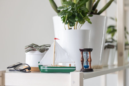 A charming green and white model sailboat stands prominently on a white shelf, flanked by various potted houseplants including one with fuzzy leaves and a larger leafy plant, alongside a classic wooden hourglass with blue sand and a vibrant green watering can in the background, all contributing to a peaceful home decor setting.