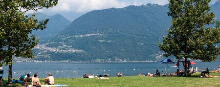 Lake Como, Italy - July 21, 2019: People resting on beach near lake. Trees and Alp mountains on background. Panoramic view.のeditorial素材