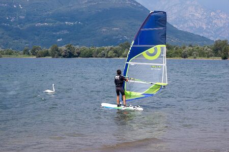 Lake Como, Italy - July 21, 2019. Water sport: windsurfer riding on a bright sunny summer day near the Colico, town in Italy, and swimming swan. Alp mountains on background. Man and bird. People and nature.のeditorial素材
