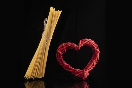 Wicker red heart and flying bunch of raw dry spaghetti on black background isolated. Italian pasta, macaroni. Horizontalの写真素材