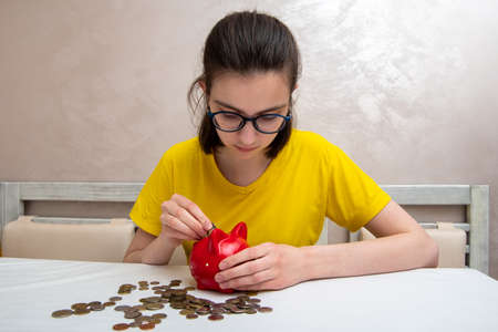 young girl put money coins in a red piggy bank to save money, several coins on the table.の写真素材