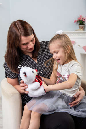 Mother and daughter playing with a teddy bear on a gray background. Daughter sits on mom lap. Family traditions. Happy family.の写真素材