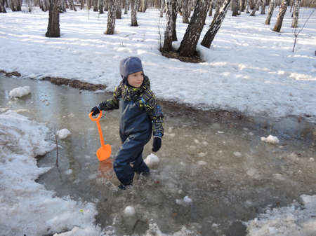 A child in blue waterproof pants, a jacket and a hat stands in a deep puddle. sunny day.の写真素材
