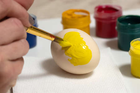 painting eggs in yellow shows a hand with a brush. Nearby are open multi-colored paints. selective focus.の写真素材