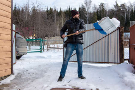 Caucasian man with a beard in a black jacket, blue jeans and a black hat cleans snow in a parking lot for a car.の写真素材