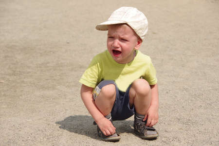 Caucasian boy in a yellow T-shirt, dark shorts, a light cap sits on the road and cries. Family...