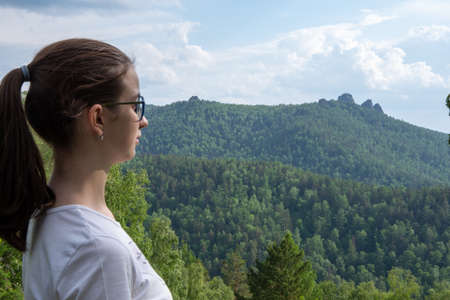 A lonely young traveler girl in a white T-shirt looks at the mountains, the trees, the forest. Outdoor recreation concept.の写真素材