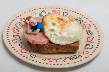 creative work: a boy lies on a toast bread with an egg and sausage on a plate on a light background.の写真素材
