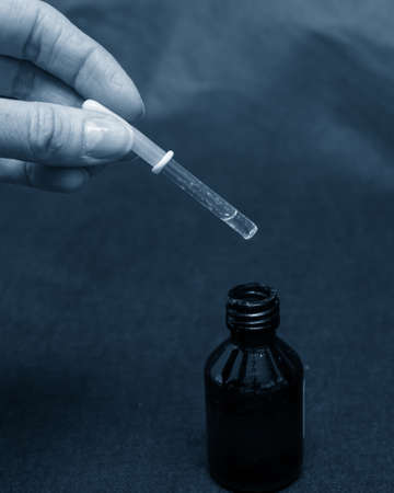 A hand holds a glass dropper with a yellow rubber tip with liquid over a dark brown bottle, close-up, tinted in blue trend 2020.の写真素材