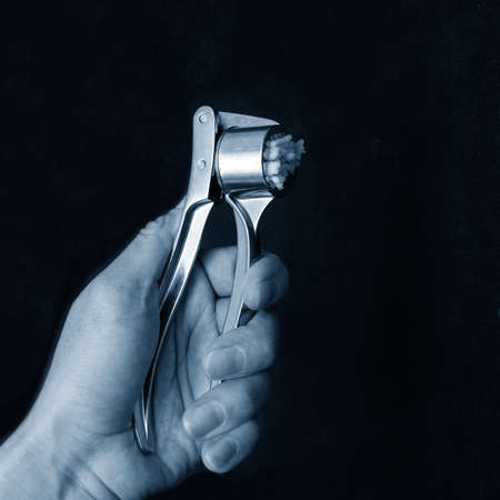 Hand of a woman cook squeezes garlic in a metal squeezer on a black background, close-up, classic blue, fashion color, color 2020の写真素材