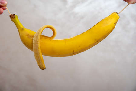 Hands holding peeled banana by wooden sticks, close-up, over gray background.