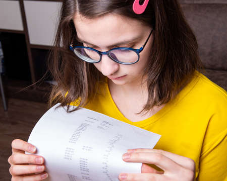 Portrait of a beautiful serious Caucasian girl in a yellow T-shirt looks at a printed report, work...