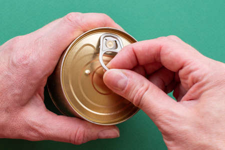 The hand of an adult holds a can of canned food; the second hand pulls the key, opens the can. Top view on green background with copy space. Close-upの写真素材