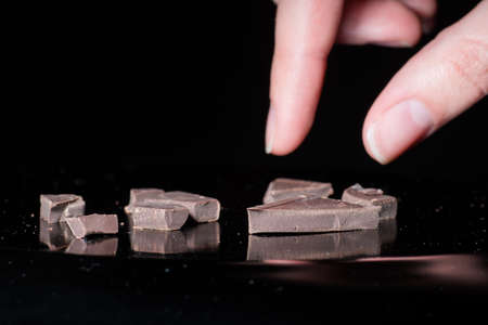 Close-up female hand takes pieces of black bitter chocolate isolated on black background, low key.の写真素材
