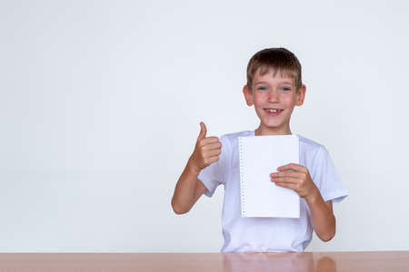 Portrait of a cute caucasian boy holding a white notebook in his hand, showing thumbs up. Boy at a...