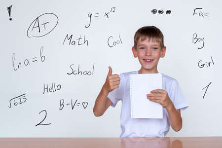 Portrait of a cute caucasian boy holding a white notebook in his hand, showing thumbs up. Boy at a...